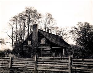 A two story log cabin with a stone chimney behind a rustic log fence. 